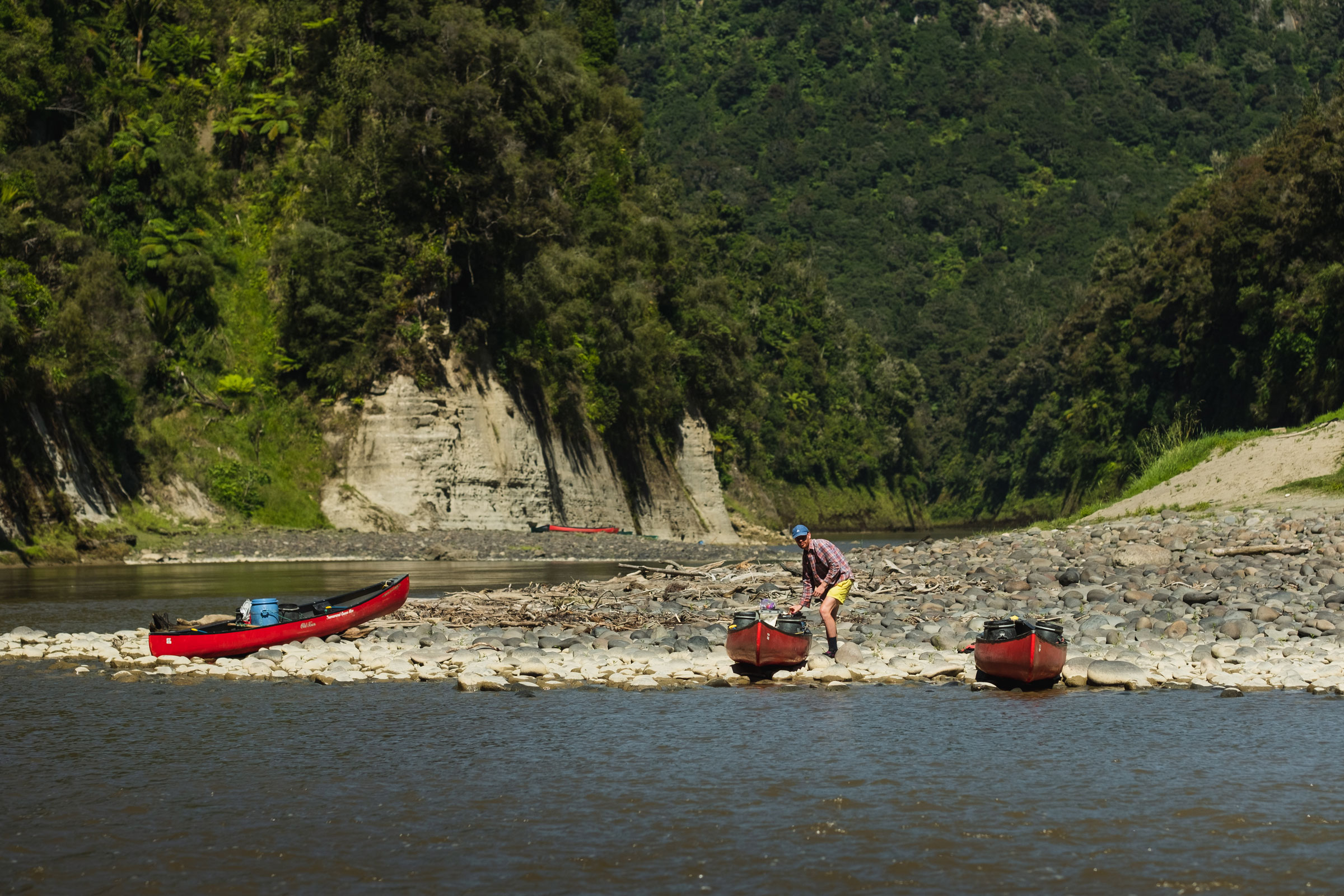 A man is standing on the rocky banks of the Whanganui River with three red canoes near the bank